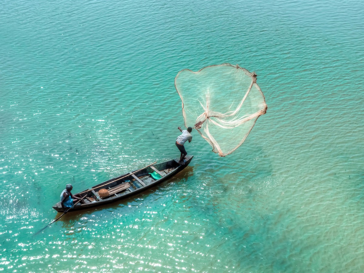 A fisherman casting a net from a small boat on clear green water