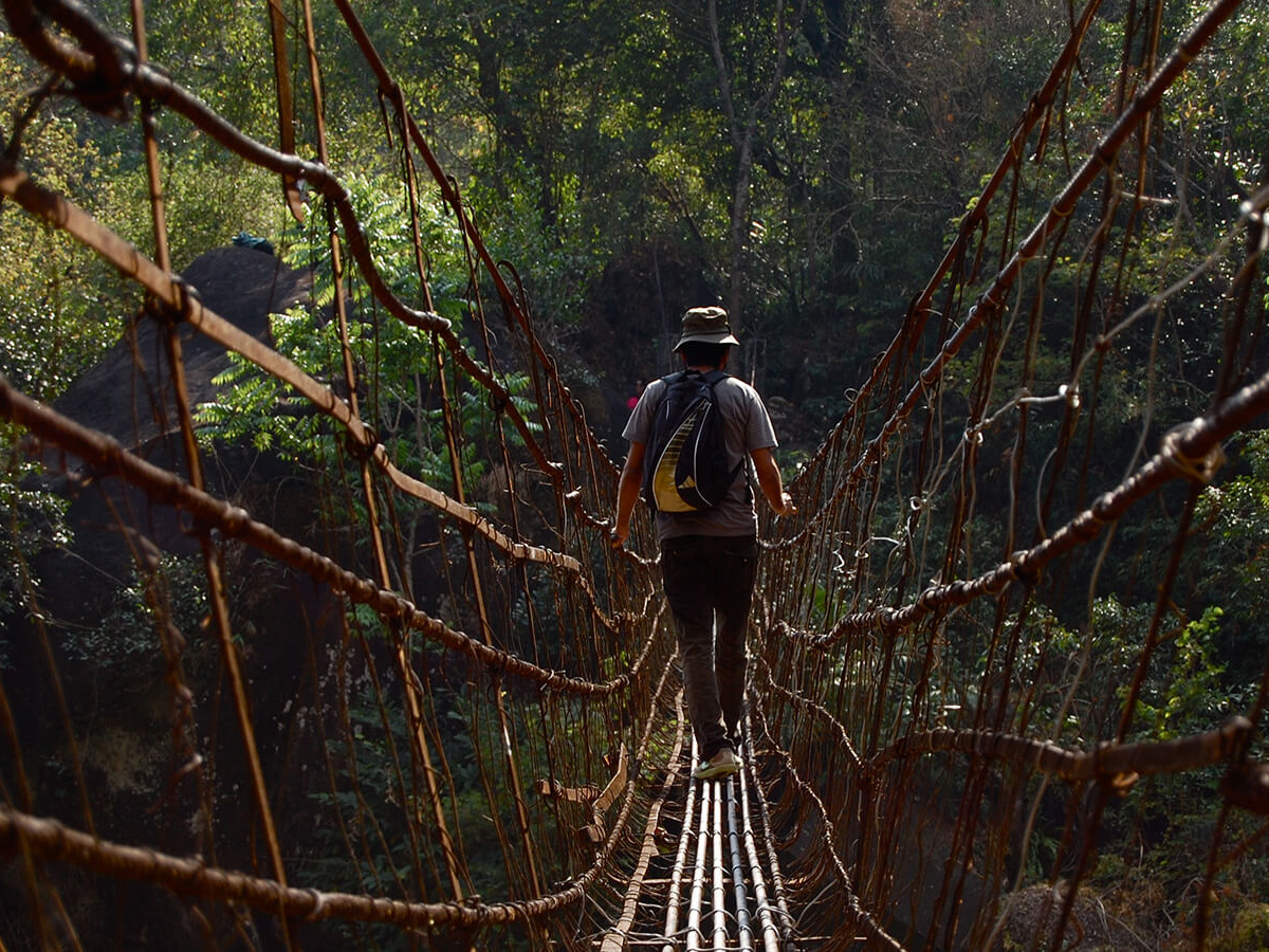 A hiker crossing a long cane and wire suspension bridge in the forest