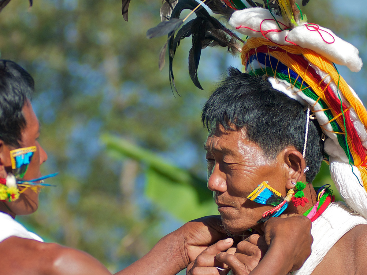 Two Naga men in feathered headdresses, one painting the other’s face