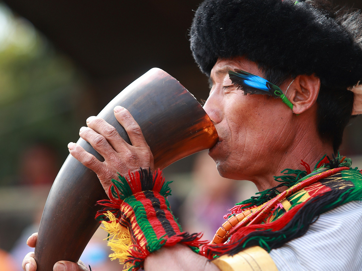 A Naga man in a feathered headdress blowing a buffalo horn