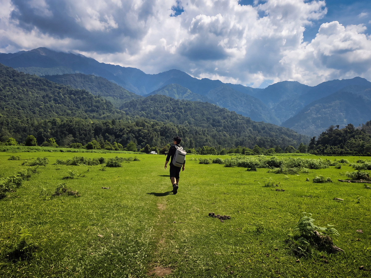 A trekker walking across a green meadow below forested hills