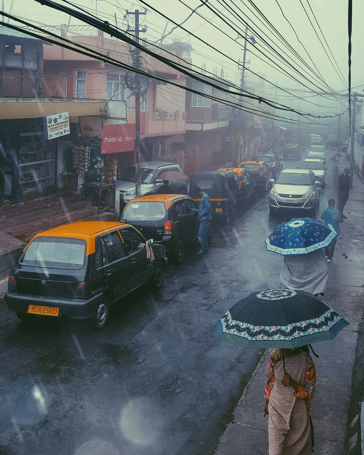 Taxis and people with umbrellas on a rainy Shillong street under tangled wires