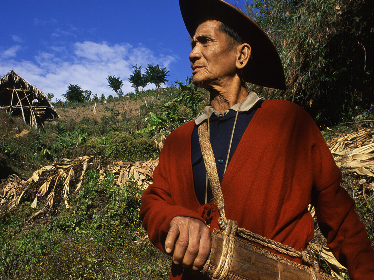 An elder in a red coat and cane hat beside a hill farm