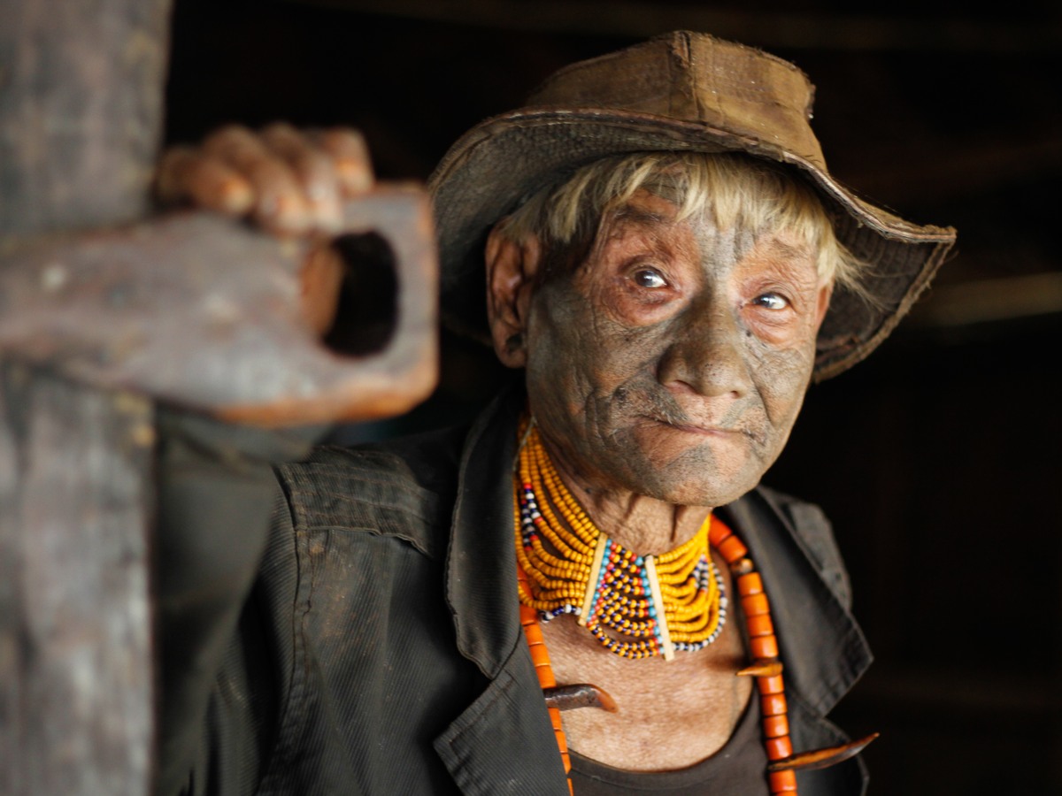 A tattooed Konyak elder in a worn hat and orange bead necklaces, inside the morung