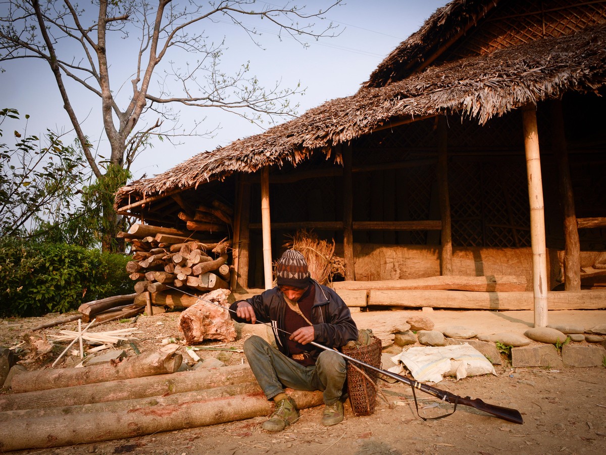 A man working on a gun barrel outside a thatched Konyak longhouse