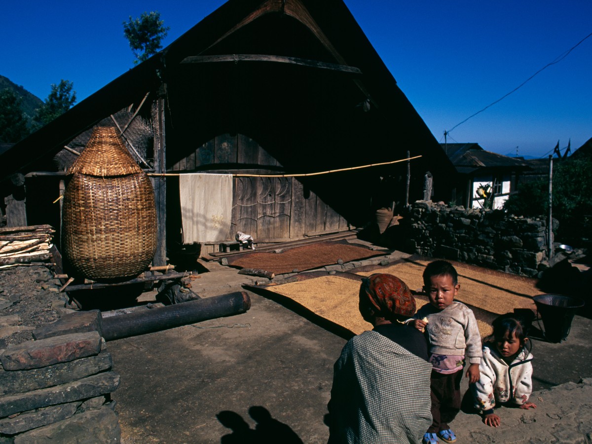 Rice drying on mats outside a Naga house, a woman and two children beside a tall cane basket